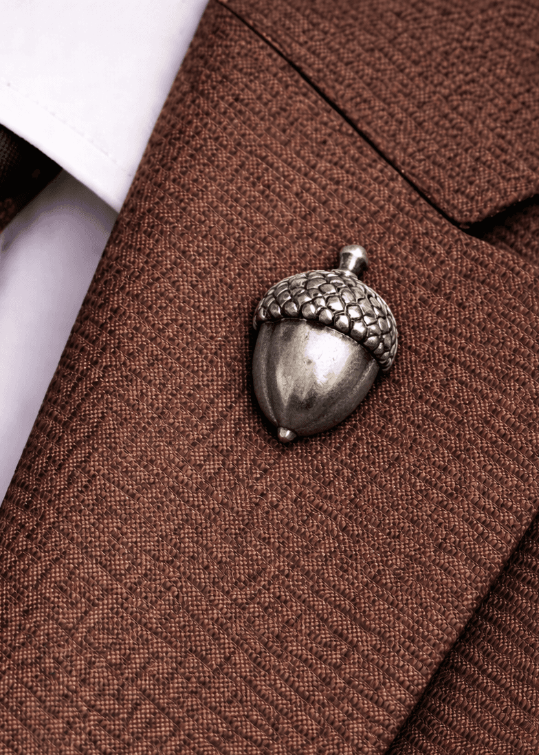The acorn pin in antique nickel, worn on a rust-brown lapel over a white shirt