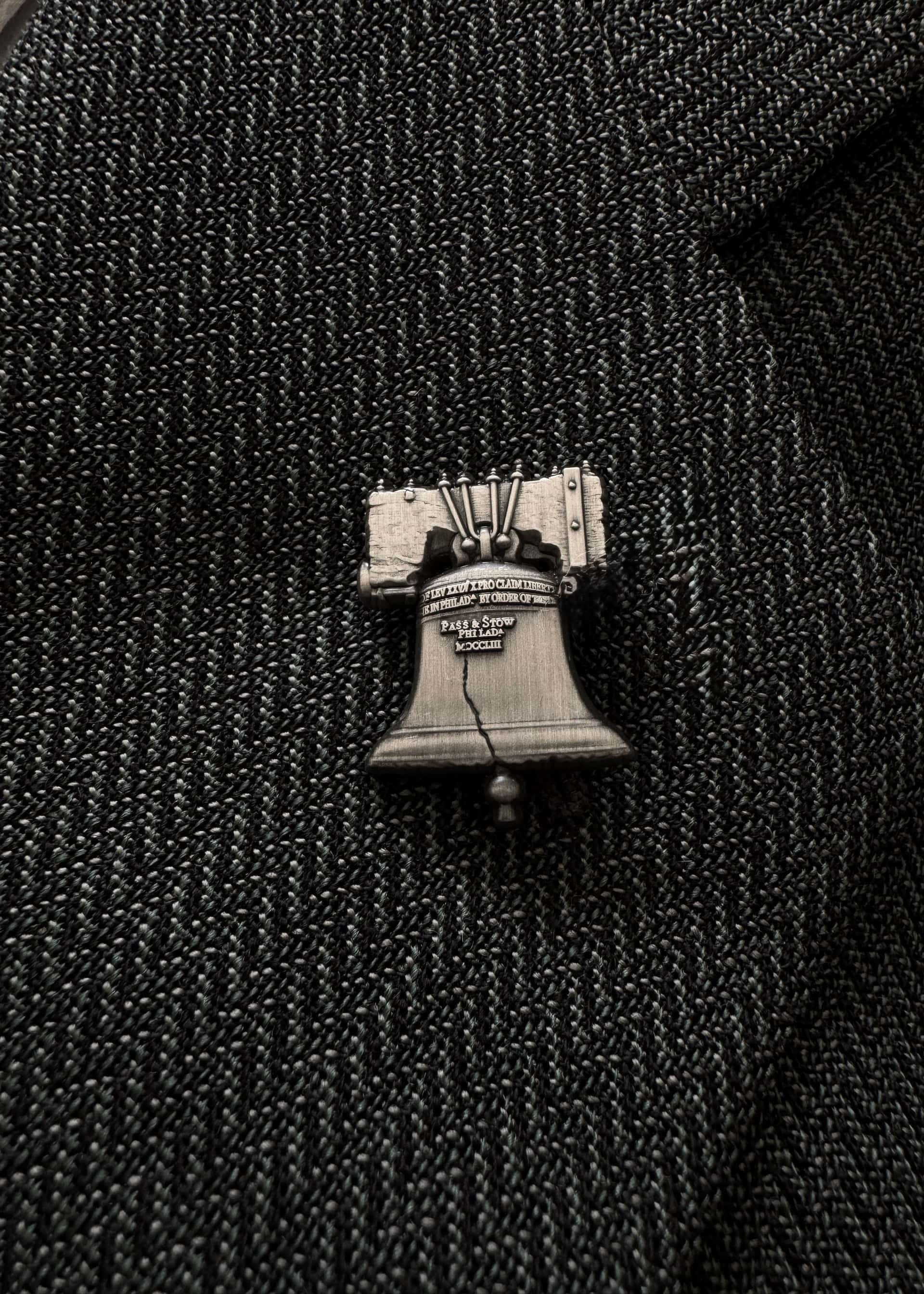 The Liberty Bell pin in antique nickel, worn on a dark herringbone lapel